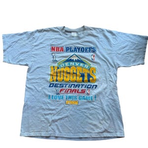 May include: Light gray t-shirt featuring the text "NBA Playoffs DENVER NUGGETS DESTINATION FINALS I LOVE THIS GAME! 2005" in blue, yellow, and red. The shirt has a crew neck and short sleeves.