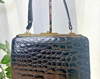 May include: A vintage black handbag with a textured, crocodile-like pattern. The rectangular bag features a gold-toned clasp and handle attachments. The handle is a matching black color. The bag is displayed on a wooden surface.