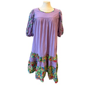 May include: A lavender midi dress featuring a round neckline, puffed sleeves with crisscross ribbon detailing, and a tiered skirt. The sleeves and the two bottom tiers of the skirt are accented with a vibrant, multi-colored floral pattern.