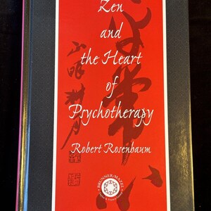 Pode incluir: Uma capa de livro vermelha com o título "Zen e o coração da psicoterapia" de Robert Rosenbaum. O título está escrito em uma fonte estilizada com caracteres japoneses no fundo.