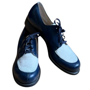 May include: A pair of two-tone oxford shoes. The shoes feature a navy blue leather upper with a light gray suede toe cap. The shoes have dark blue laces and a black sole. The shoes are displayed on a white background.