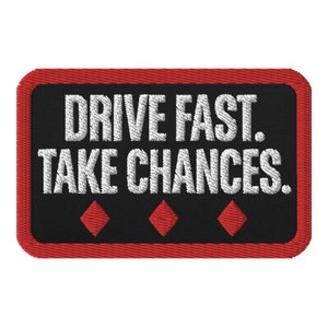 Op de afbeelding: Zwarte en rode geborduurde patch met witte tekst die "DRIVE FAST. TAKE CHANCES." luidt en drie rode ruiten.