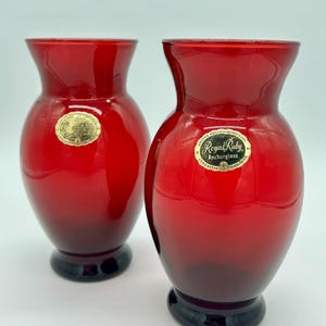 May include: Two ruby red glass vases with a rounded body and flared rim. Each vase has a black base and a gold label that reads "Royal Ruby Anchorglass". These vintage vases are suitable for floral arrangements.