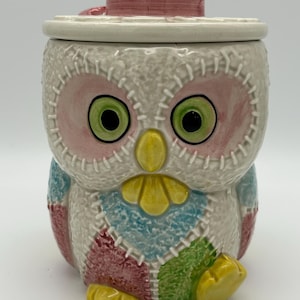 May include: A ceramic owl cookie jar with a patchwork design in pink, blue, green, and yellow. The owl is wearing a pink hat with a white brim and a pink ribbon. The jar has a lid.