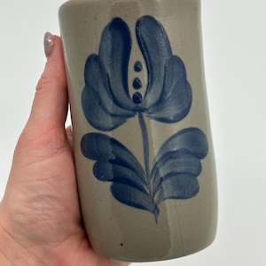 May include: A cylindrical ceramic mug with a beige background and a large blue floral design. The flower has multiple petals and a stem with leaves. The mug is held in a hand.