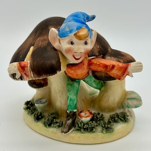 Peut inclure: Une figurine en céramique d'un lutin souriant portant un chapeau bleu et des vêtements rouge et orange, debout sur un champignon avec des détails bruns et blancs. La figurine est entourée de verdure et de fleurs.
