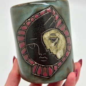 May include: A cylindrical ceramic mug with a teal and blue glazed finish. The front features a relief design of a stylised face split into a black side and a textured cream side, surrounded by a border of small, rectangular, dark pink segments.