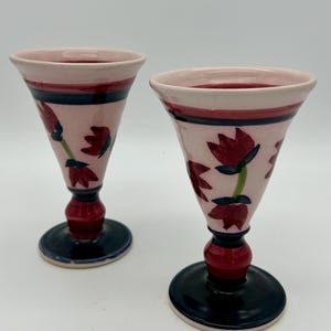 May include: Two pink ceramic goblets with a flared top, decorated with red floral designs and green stems. The goblets have a dark blue base and a red stem. The rim of the goblets is a darker pink.
