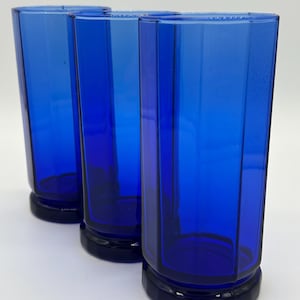 May include: Three tall, cobalt blue drinking glasses. Each glass has a faceted design and a dark blue base. The glasses are made of a transparent material. The glasses are arranged in a row, with a white background.