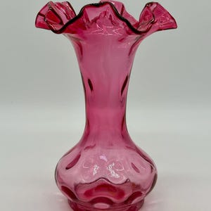 May include: A pink glass vase with a ruffled rim and a bulbous base. The vase has a slender neck and a flared top, with a dark trim outlining the ruffled edges. The glass has a glossy finish, reflecting light.