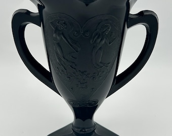 Peut inclure: Un vase en verre noir avec deux anses et une base carrée. Le vase est décoré d'un motif en relief de deux femmes.