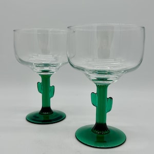 Op de afbeelding: Twee heldere cocktailglazen met groene cactusvormige stelen. De glazen hebben een afgeronde kom en een brede voet. De cactusstelen zijn felgroen en geven het glaswerk een unieke uitstraling.