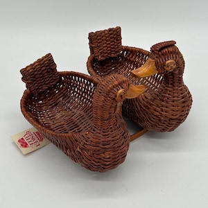 May include: Two brown woven wicker baskets shaped like ducks, featuring smooth wooden beaks and a rectangular tag attached to one side that reads BASKET VILLE. The baskets are connected at the base and have open, hollow centres for storage.