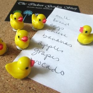 May include: Five yellow rubber ducky magnets with red beaks and black eyes are holding a white piece of paper with a handwritten grocery list that says "milk, bread, eggs, cheese, bananas, apples, grapes, avocado".