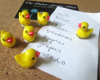 May include: Five yellow rubber ducky magnets with red beaks and black eyes are holding a white piece of paper with a handwritten grocery list that says "milk, bread, eggs, cheese, bananas, apples, grapes, avocado".
