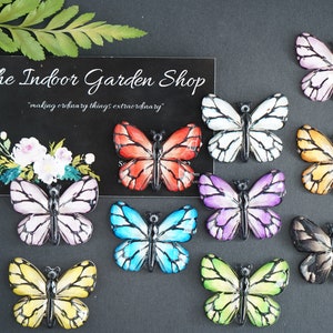 May include: Ten colorful butterfly embellishments with black outlines. The butterflies are made of resin and have a glossy finish. The butterflies are arranged in a row on a black background. The text "The Indoor Garden Shop" is visible in the background.