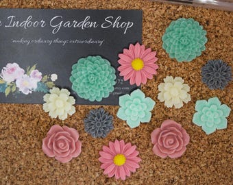 May include: A collection of colorful, decorative flower-shaped items in various sizes and colors, including pink, green, and white. The flowers are arranged on a corkboard surface, with a business card in the background.