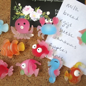 May include: A set of colorful sea creature push pins, including a blue crab, a pink octopus, a green fish, a red fish, a blue fish, a yellow fish, a pink fish, and a blue seahorse. The push pins are on a corkboard with a white note that says "Milk, bread, eggs, cheese, bananas, apples, limes".