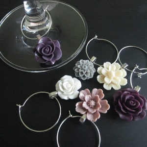 May include: Six different flower-shaped wine glass charms. The charms are made of metal and have a variety of colours, including purple, white, grey, cream, and pink.