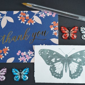 May include: A thank you card with a floral pattern and gold lettering. The card is surrounded by five decorative butterflies in various colors, including red, white, blue, and pink. The butterflies are arranged in a semi-circle around the card.