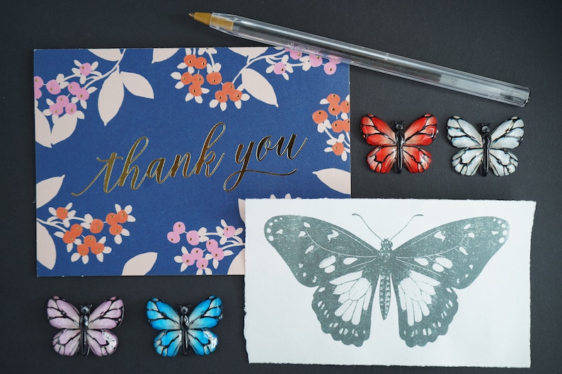 May include: A thank you card with a floral pattern and gold lettering. The card is surrounded by five decorative butterflies in various colors, including red, white, blue, and pink. The butterflies are arranged in a semi-circle around the card.