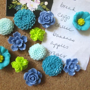 May include: A corkboard with a variety of blue, green and yellow flower-shaped push pins. A piece of paper with the words "bread, eggs, cheese, banana, apples, grapes" is pinned to the corkboard.
