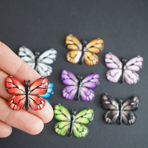 May include: Assortment of colorful butterfly-shaped embellishments. The butterflies are made of resin and have a glossy finish. The colors include red, orange, yellow, green, blue, purple, white, and black.