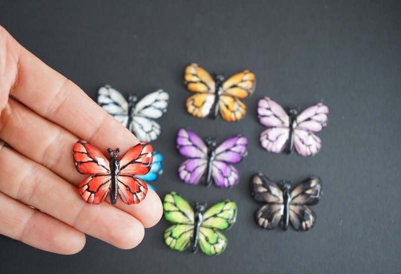 May include: Assortment of colorful butterfly-shaped embellishments. The butterflies are made of resin and have a glossy finish. The colors include red, orange, yellow, green, blue, purple, white, and black.