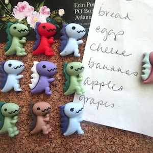 May include: A collection of colorful dinosaur-shaped magnets in various colors, including green, red, blue, and purple. A handwritten grocery list is attached to a corkboard. The magnets are small and decorative.