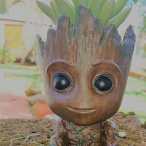 Puede incluir: Un macetero de Groot de madera marrón con una planta suculenta verde que crece en la parte superior. La figura sostiene un pequeño nido de pájaro con dos pájaros amarillos dentro.