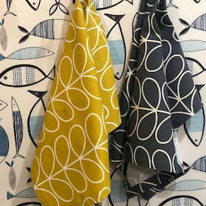 Könnte beinhalten: Zwei Küchentücher hängen an einer Wand mit Fischmuster. Ein Tuch ist senfgelb mit weißem Blumendesign, das andere dunkelgrau mit weißem Blumendesign. Die Wand hat ein sich wiederholendes Muster aus blauen und schwarzen Fischen.