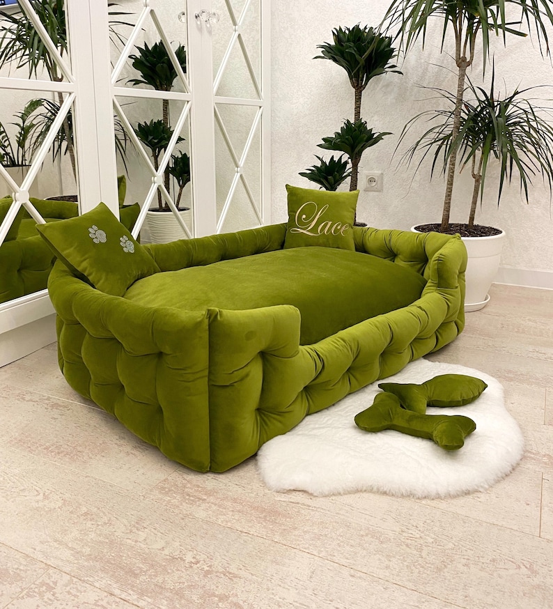 May include: A green velvet dog bed with a tufted design, two matching pillows, and two bone-shaped toys. The bed sits on a white faux fur rug. The word "Lace" is embroidered on one pillow.
