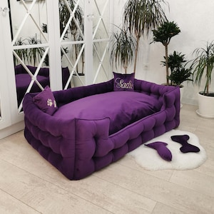 May include: A plush, deep purple dog bed with a tufted design. It features a rectangular shape with a soft, cushioned interior and two matching throw pillows. One pillow has a paw print, and the other has the name "Sadie" embroidered on it.