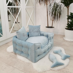 May include: A light blue, tufted pet bed with two matching pillows and two bone-shaped toys. The bed sits on a white, fluffy rug. One pillow has a paw print design, and the other has the name "Rocky" embroidered on it.
