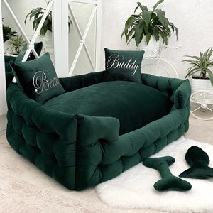 May include: A dark green velvet dog bed with a tufted design. The bed has two pillows with the names "Buddy" and "Beau" embroidered on them. The bed is on a white fluffy rug.