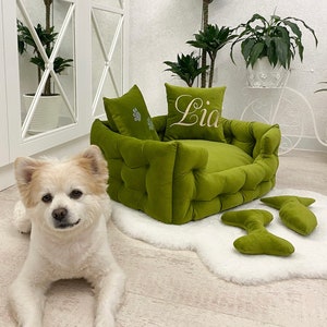 May include: A green, tufted pet bed with two matching pillows, one embroidered with the name "Lia" in white script. Two bone-shaped toys sit on a white faux fur rug. A small, white and tan dog is lying on the floor.