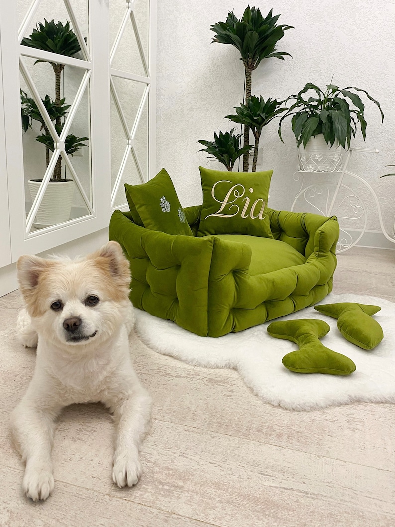 May include: A green, tufted pet bed with two matching pillows, one embroidered with the name "Lia" in white script. Two bone-shaped toys sit on a white faux fur rug. A small, white and tan dog is lying on the floor.