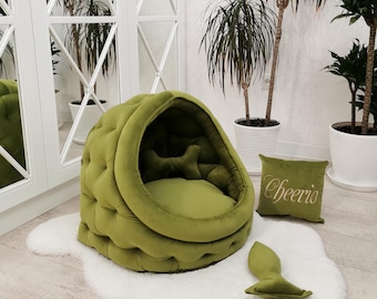 May include: A tufted olive green pet bed with a dome-shaped canopy, a removable round cushion, and a bone-shaped pillow inside. Included is a square olive green pillow with the word Cheerio embroidered in gold and a small bone-shaped toy.