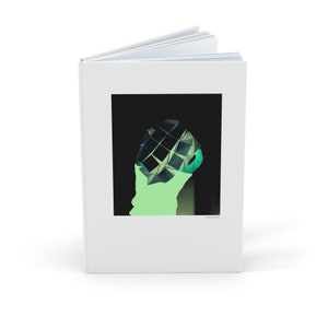 Op de afbeelding: Een open wit notitieboek met een abstracte kunstprint in zwart en groen. Het kunstwerk heeft een geometrisch ontwerp tegen een donkere achtergrond, met een neon groen silhouet. Het notitieboek is open en toont lege pagina's.