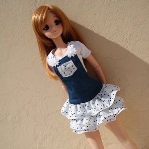 May include: A doll with long brown hair wearing a blue denim overall dress with a white ruffle skirt with black polka dots.