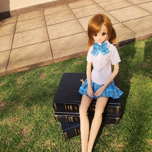 May include: A doll wearing a white shirt with a blue bow tie, a blue and white plaid skirt, and white socks. The doll is sitting on a stack of three black books with gold lettering on the spines. The doll is sitting on a green grassy surface.
