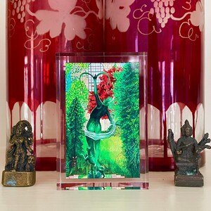 May include: A clear acrylic display case with a colorful painting of a woman standing in a green forest. The woman is wearing a green dress and has her arms raised above her head. The painting is set against a background of green trees and a red sky.