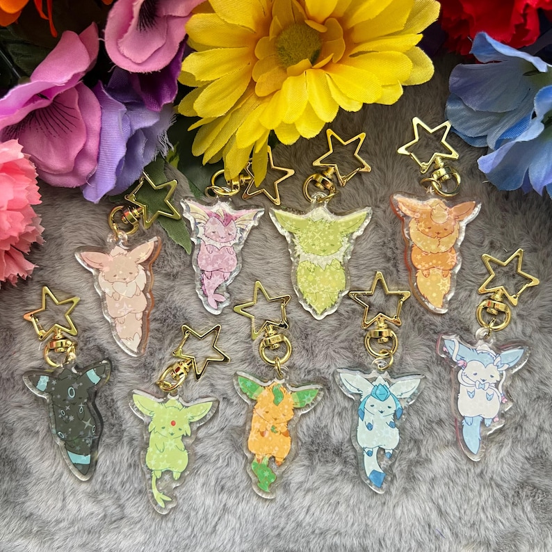May include: Nine colorful keychains featuring different Pokemon characters. Each keychain is made of clear acrylic with a gold star-shaped ring. The characters are depicted in a cartoon style and are surrounded by flowers.
