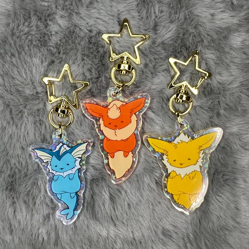 May include: Three iridescent keychains featuring cartoon versions of the Pokemon Eevee evolutions: Vaporeon, Flareon, and Jolteon. Each keychain is attached to a gold star-shaped swivel clasp.
