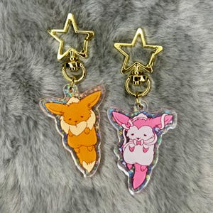 May include: Two gold star-shaped keychains with clear acrylic charms. One charm features a pink Eevee with a blue bow, and the other charm features an orange Eevee with a yellow bow.