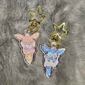 May include: Two iridescent keychains featuring cartoon characters. The keychains are attached to gold star-shaped charms with a gold swivel clasp. The characters are a pink and white fox and a blue and white fox.