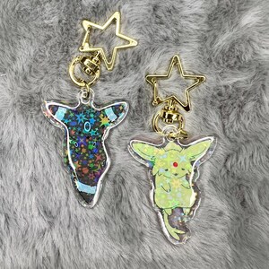 May include: Two iridescent holographic keychains featuring Pokemon characters. The keychain on the left features a black and green Umbreon with a gold star charm. The keychain on the right features a green and yellow Espeon with a gold star charm.