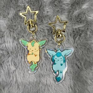 May include: Two gold keychains with clear acrylic charms featuring cartoon animal characters. The charms are iridescent and have a holographic effect. The first charm is a light green and yellow creature with a green leaf tail. The second charm is a light blue creature with a blue tail.