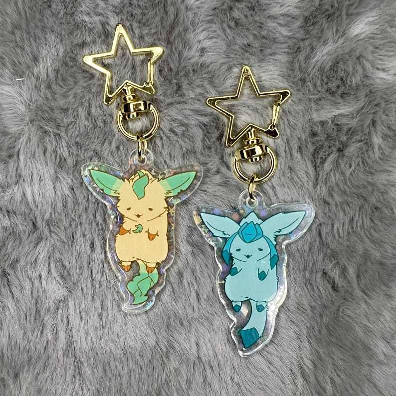 May include: Two gold keychains with clear acrylic charms featuring cartoon animal characters. The charms are iridescent and have a holographic effect. The first charm is a light green and yellow creature with a green leaf tail. The second charm is a light blue creature with a blue tail.