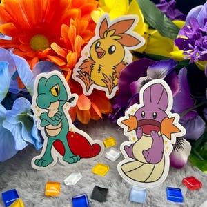 May include: Three cartoon Pokemon stickers. A green and red lizard-like Pokemon with a yellow eye, a yellow bird-like Pokemon with orange flames, and a purple Pokemon with orange fins.